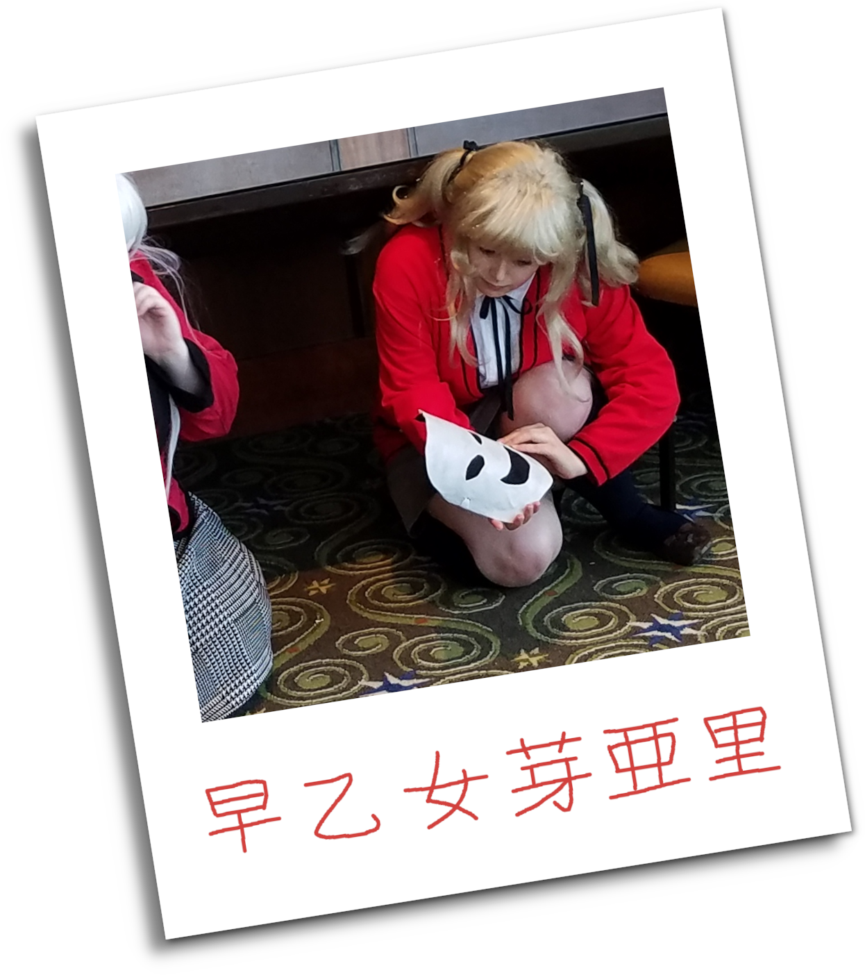 A young woman cosplays Mary Saotome from from Kakegurui. She has a blonde wig worn in pigtails tied with black ribbons, and wears a red cardigan, a white button up with a ribbon at the neckline ,a grey skirt, blue knee-high stockings, and brown shoes. She kneels on the ground of a formal room, picking up Ririka's mask. A woman cosplaying Ririka has been partially cropped out of the photo to protect her identity. The original image has been framed with a polaroid, which is signed at the bottom. The Kanji '早乙女芽亜里' (Mary Saotome in English) are written in red pen, to match with Mary's school uniform.